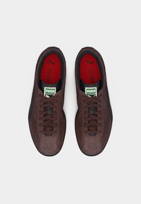 Ζεύγος καφέ αθλητικών παπουτσιών Puma με μαύρες σόλες, καφέ κορδόνια, κόκκινες εσωτερικές σόλες Ortholite και πράσινες ετικέτες με το λογότυπο, βλέπονται από πάνω σε λευκό φόντο.