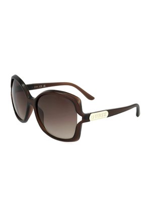 Occhiali da sole oversize marroni con lenti sfumate e logo "GUESS" dorato sulle aste, vista angolata su sfondo bianco.