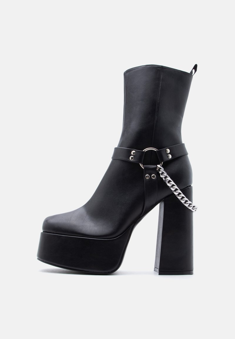 Μαύρο μποτάκι πλατφόρμας με χοντρό ψηλό τακούνι, ασημένια αλυσίδα και λεπτομέρεια με λουρί σε απλό ανοιχτό φόντο.