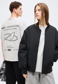 Νεαρός άνδρας με λευκό μπλουζάκι με γραφικό μοιάζει προς τα αριστερά, γυναίκα με μαύρο μπουφάν τύπου bomber και γκρι παντελόνι κοιτάζει προς τα δεξιά σε απλό λευκό φόντο.