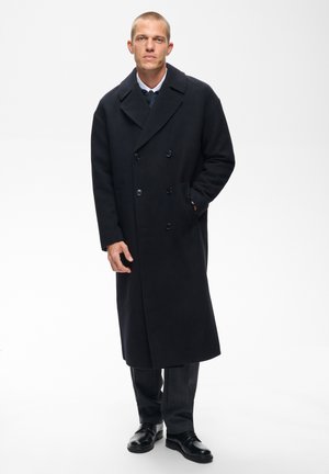 Man standing wearing a long black double-breasted coat, dark trousers, and black shoes on a white background.