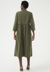 Olijfgroen jurk met driekwartmouwen, een versmallende taille en een lengte tot op de kuit. Gemaakt van zachte, lichte stof en heeft een hoge hals.