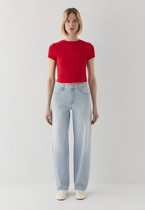 Frau mit kurzen blonden Haaren, die ein rotes, kurzes T-Shirt, hellblaue Jeans mit hoher Taille und weiße Schuhe trägt, stehend vor weißem Hintergrund.