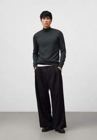 Maglione a collo alto verde scuro, design aderente, texture lavorata a maglia. Pantaloni neri a vita alta con pieghe, vestibilità ampia, tessuto liscio, indossati con scarpe nere.