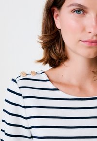 Top a righe bianco e blu navy, dotato di un collo rotondo e dettagli texturizzati sulle spalle in beige, con due bottoni circolari sulla spalla sinistra.