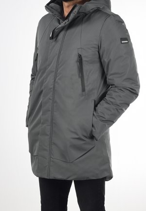 Homme portant un manteau d'hiver gris à capuche avec poches poitrine zippées et mains dans les poches latérales, debout sur fond blanc.