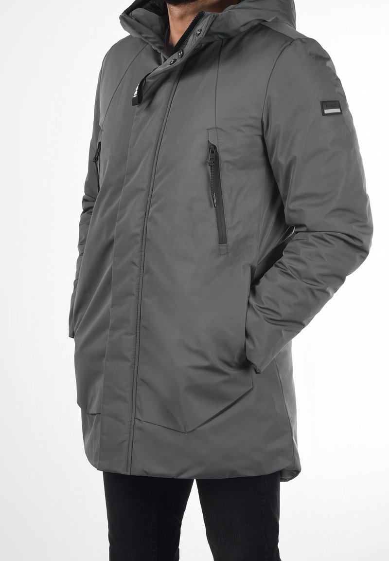 Homme portant un manteau d'hiver gris à capuche avec poches poitrine zippées et mains dans les poches latérales, debout sur fond blanc.
