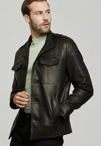 Zwarte leren jas met een gestructureerde afwerking, knoopsluiting, twee borstzakken en een kraagontwerp, gedragen over een lichtgroene trui.