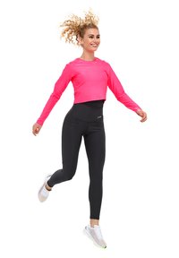 Top cropped a maniche lunghe rosa abbinato a leggings neri a vita alta, con texture liscia e vestibilità atletica, completato da scarpe bianche.