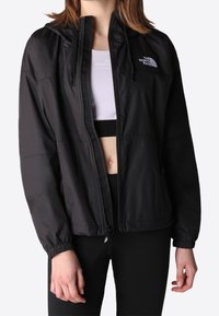 Giacca a vento nera con zip frontale, cappuccio regolabile e polsini elasticizzati. Presenta un dettaglio con logo sul petto e tessuto leggero.