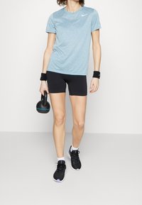 Jasnoniebieska sportowa koszulka z krótkim rękawem, czarne teksturowane szorty, czarne trampki z białymi akcentami, trzyma czarny kettlebell z niebieskim detalem.