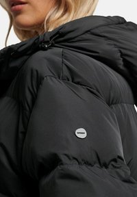 Sort pufferjakke med en skinnende, polstret tekstur. Har en hætte med snor og et lille gråt logo-patch på venstre ærme.