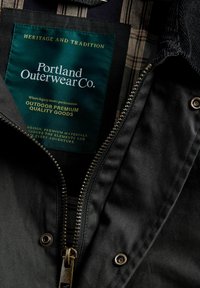 Črna jakna za zunanjo oblačilo z zeleno oznako, ki pravi "Portland Outerwear Co", z zadrgo, gumbnimi poudarki in karo podlogo.