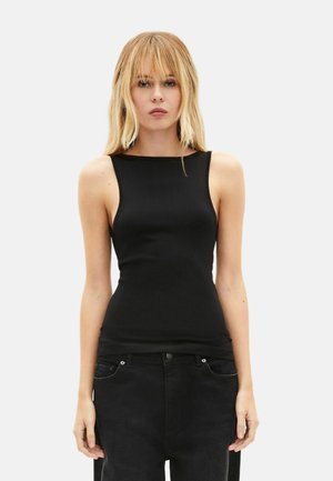 Jeune femme aux cheveux blonds et raides, portant un haut noir sans manches et un pantalon noir, debout devant un fond blanc uni.