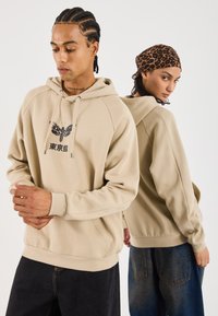Beige hoodie med kængurulomme, der har et grafisk design og snor i hætten. Modellerne bærer denimbukser; en af dem har et leopardmønstret tørklæde.