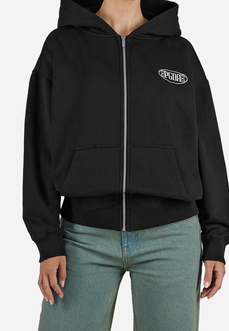 Personne portant un sweat à capuche noir zippé avec un petit logo blanc sur la poitrine et un jean bleu clair, visible du milieu du cou aux hanches.