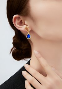 Pendientes de oro con una gema azul en forma de lágrima rodeada por un halo de pequeñas piedras claras, con un diseño de eslabones texturizados.