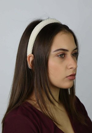 Hoofdband van crèmekleurige stof met een gladde textuur. Het bandje is licht gevoerd en heeft een afgeronde vorm, gedragen door een model met lang haar.