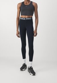 Crop top Nike Pro w ciemnoszarym kolorze, połączony z granatowymi legginsami z białym elastycznym pasem. Modelka nosi czarne obuwie sportowe i białe skarpetki.