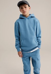 Felpa leggera blu realizzata in materiale morbido, con tasca frontale e polsini a coste. Indossata con pantaloni della tuta coordinati e una maglietta bianca sotto.