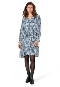 Abito blu a fantasie con maniche lunghe, scollatura con bottoni e polsini arricciati. Abbinato a stivali neri alla caviglia e collant neri trasparenti.