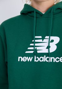 Sweat à capuche vert avec cordon de serrage, présentant un grand logo blanc "NB" et le texte "new balance" en dessous, fabriqué en tissu doux et texturé.