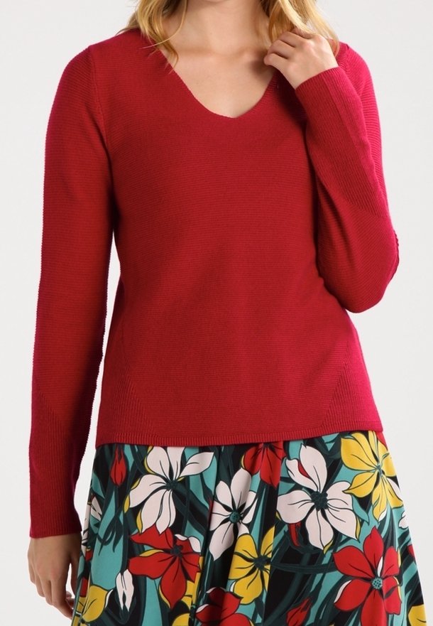 Femme portant un pull rouge à col en V et manches longues, ainsi qu'une jupe fleurie avec des fleurs rouges, blanches et jaunes sur fond vert.