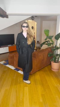 Μαύρο διπλόμπουτο παλτό με ζώνη σε δεσίματα, φορεμένο πάνω από ανοιχτό ροζ μπλουζάκι. Σκούρα τζιν και μπεζ τσόκαρα. Μακριά μαλλιά και μαύρα γυαλιά ηλίου.