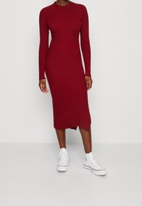 Femme vêtue d'une robe midi ajustée à manches longues côtelées de couleur rouge foncé, associée à des baskets blanches montantes à plateforme, se tenant devant un fond uni.