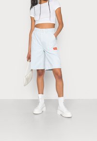 Pantalones cortos de mezclilla cargo en azul claro con un corte holgado, que cuentan con un parche de etiqueta naranja. Combinados con una camiseta corta blanca y botas gruesas blancas.
