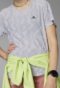 Camisa de manga corta gris claro con un patrón texturizado en zigzag, combinada con una chaqueta verde neón atada a la cintura y un reloj negro en la muñeca.