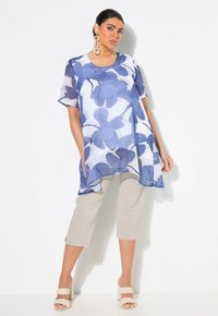 Durchsichtige blaue Blumen-Tunika mit kurzen Ärmeln, kombiniert mit beigen kaprihosen und hellen Blockabsatzsandalen. Auffälliges Blumenmuster.