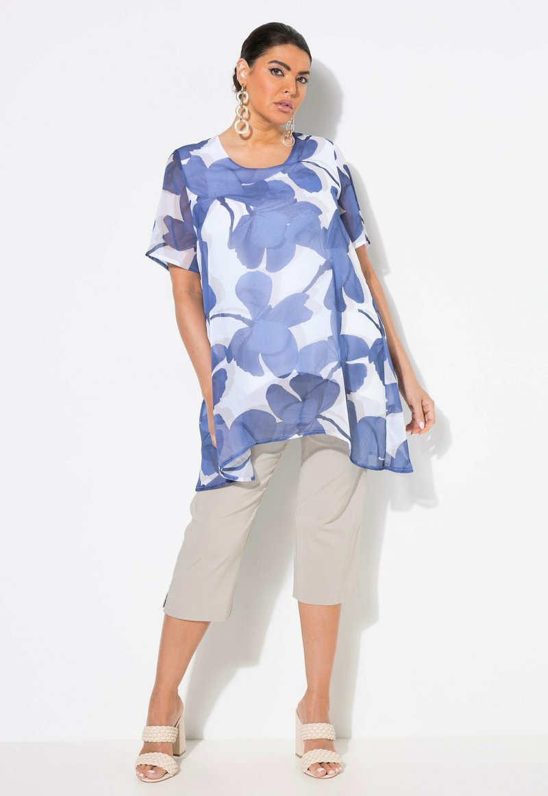 Durchsichtige blaue Blumen-Tunika mit kurzen Ärmeln, kombiniert mit beigen kaprihosen und hellen Blockabsatzsandalen. Auffälliges Blumenmuster.