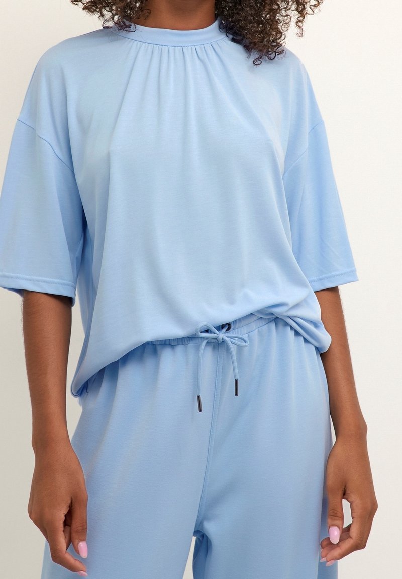 Haut bleu clair à manches courtes, coupe décontractée, avec encolure froncée, associé à un pantalon à cordon assorti sur une personne aux cheveux bouclés foncés.