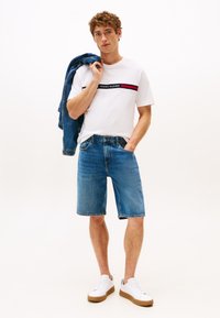 Fiatal férfi fehér Tommy Hilfiger pólóban és farmer rövidnadrágban, aki farmerdzsekit tart a vállán, az egyik kezét a zsebében tartva áll.