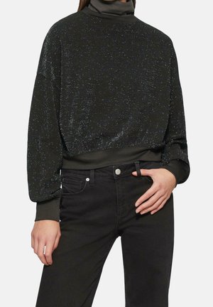 Vrouw draagt een zwart glinsterend langermig shirt met col en zwarte jeans met hoge taille, hand rust op de heup.