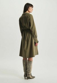 Robe chemise vert olive avec une taille cintrée, des manches longues et des poignets boutonnés ; associée à des bottes en cuir serpent jusqu'aux genoux.