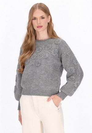Pulover gri cu broderie florală pe față, mâneci texturate și mansete ribbate. Asortat cu pantaloni de culoare deschisă.