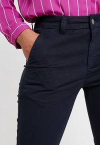 Granatowe spodnie o wąskim kroju, wykonane z gładkiej tkaniny. Posiadają przednią kieszeń, szlufki na pasek oraz metalowe zapięcie na guzik.