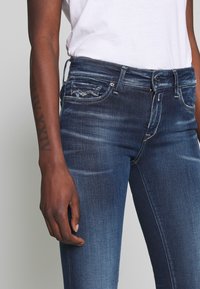 Vaqueros skinny de color azul oscuro hechos de mezclilla, con una cintura de altura media, bolsillos frontales y un leve desvanecimiento. El diseño cuenta con un clásico estilo de cinco bolsillos.