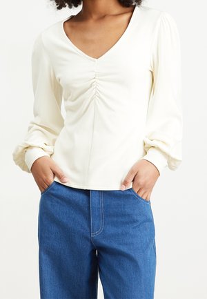 Femme portant un chemisier crème à manches longues avec un devant froncé et un pantalon en jean bleu, les mains dans les poches.