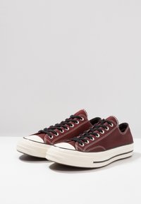 Zapatillas de lona burdeos con cordones negros, puntera y suela de goma blanca. Presenta costuras en contraste y un diseño de corte bajo.