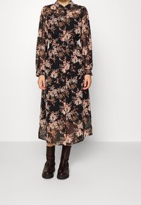 Kvinna klädd i en långärmad, svart blommig midiklänning med bälte och mörkbruna ankelstövlar, stående mot en vit bakgrund.