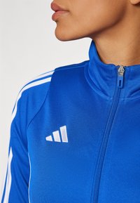 Blue athletic jacket with a high collar, featuring white stripes on the shoulders and a white logo on the chest. Zipper detail is visible.