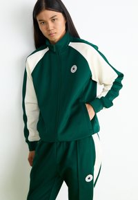 Πράσινη αθλητική φόρμα από απαλό ύφασμα, με λευκές λεπτομέρειες και στρογγυλή λογότυπο στη ζακέτα και τα παντελόνια. Ζακέτα με φερμουάρ.