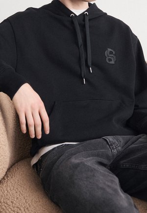 Personne assise sur une chaise beige texturée portant un sweat à capuche noir avec un logo noir sur la poitrine et un jean gris foncé.