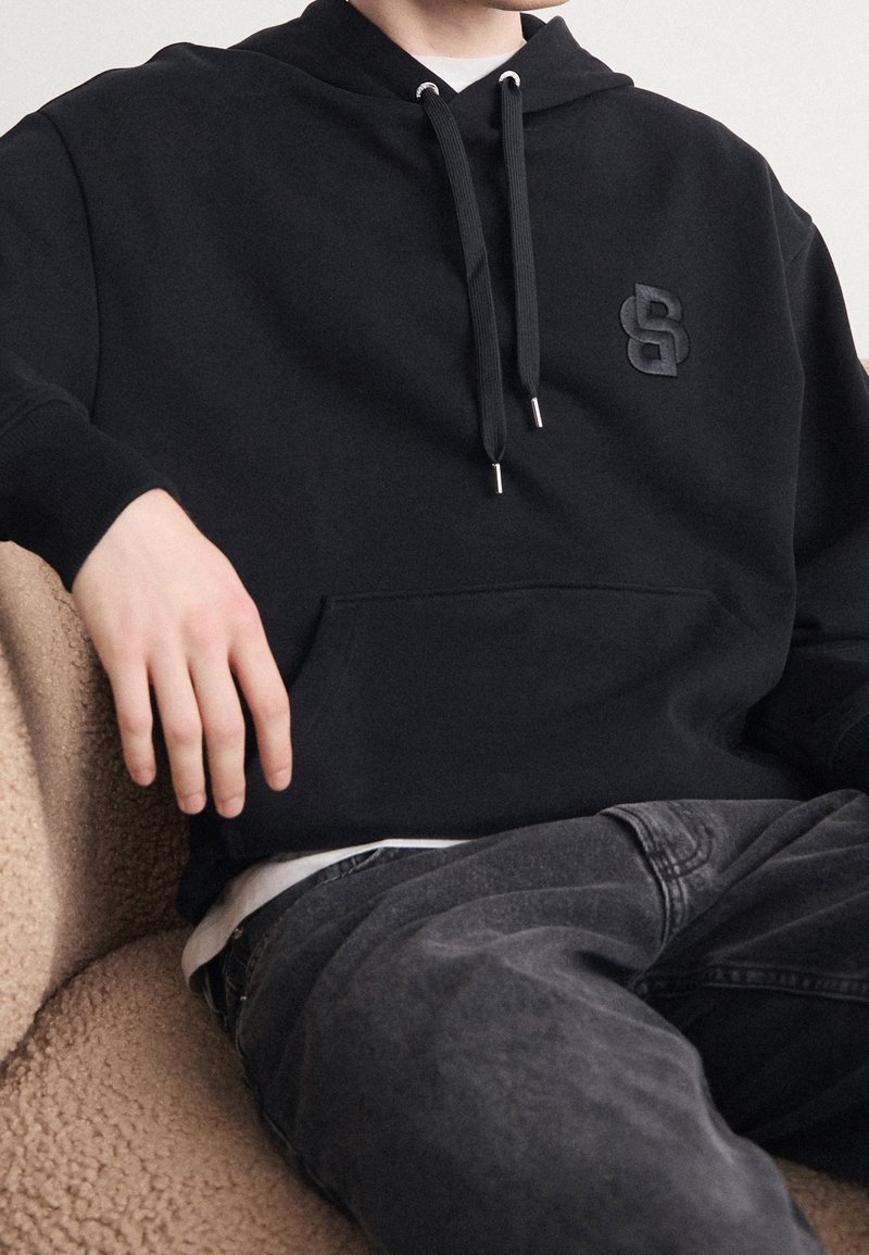 Personne assise sur une chaise beige texturée portant un sweat à capuche noir avec un logo noir sur la poitrine et un jean gris foncé.