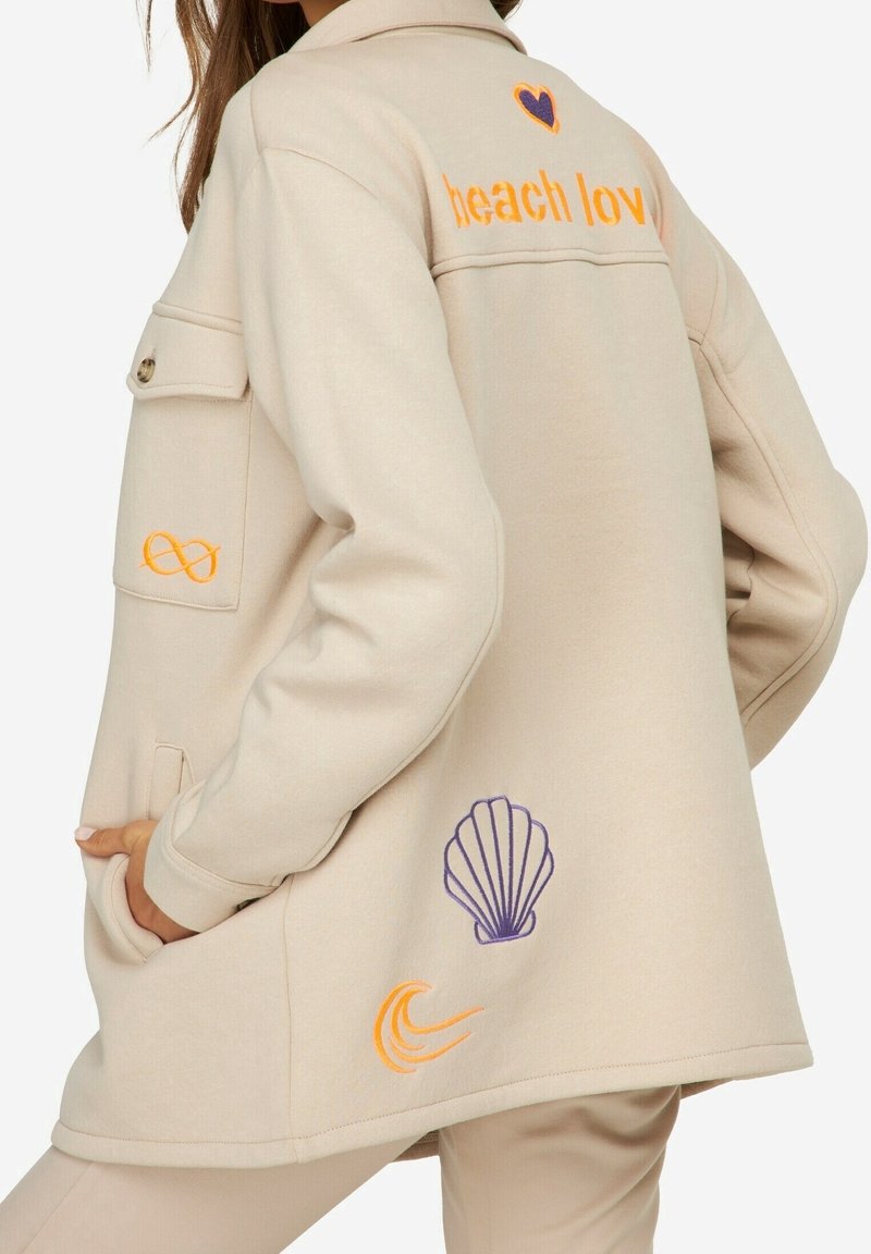Béžová bunda vyrobená z měkké tkaniny s oranžovou a fialovou výšivkou. Design zahrnuje text "beach lov", srdce, mušle a vlnu.