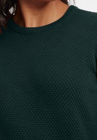 Tummanvihreä neulepaita, jossa on resorikaulus ja teksturoitu mehiläishuonemainen kuviointi. Kangas näyttää neulotulta ja pehmeältä.