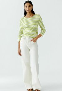 Gestreiftes grün-weißes Langarmshirt mit Rundhalsausschnitt und Knopfdetails an der Schulter, kombiniert mit weißen, weit geschnittenen Jeans und braunen Sandalen.